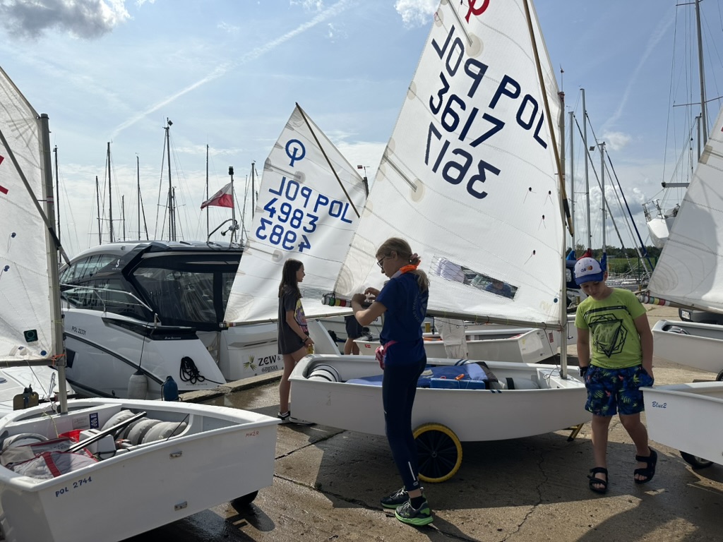 Troje dzieci przy dwóch optimistach na przyczepach w marinie
